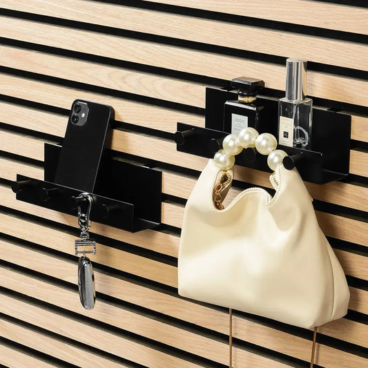 Wooden wall with black horizontal slat-like panels, holding black shelves with a phone, perfume bottles, a handbag with a pearl handle, and keys.