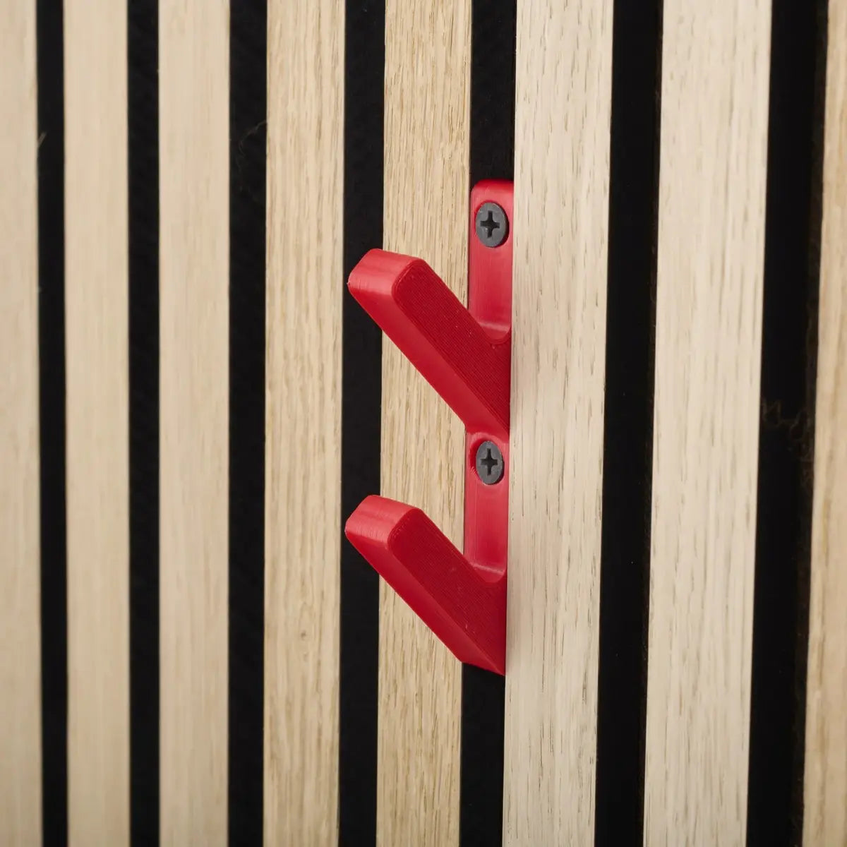 Close-up of a red double coat hook attached to a wooden slat wall with black gaps.