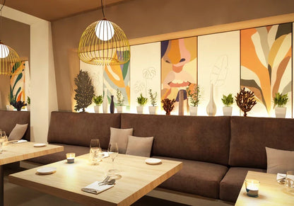 Restaurant interior with brown banquette seating, light wood tables, and modern gold pendant lighting. Artwork panels and plants decorate the space.