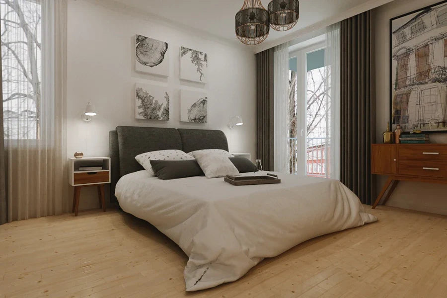 A bedroom features a gray headboard bed with white linens, flanked by a white and brown bedside table and a wooden cabinet. Artwork adorns the walls, with sheer white and brown curtains framing the windows. Overhead, a rustic chandelier adds a touch of elegance.