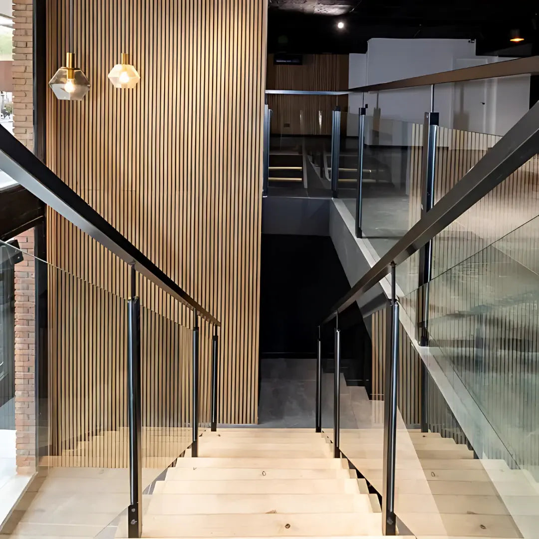 View down wooden stairs with black railings and glass panels, featuring a slatted wood wall and pendant lights.
