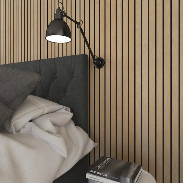Bedroom setting with a gray tufted headboard, white linens, a black adjustable wall lamp, and a stack of books on a side table against a wooden slat wall.