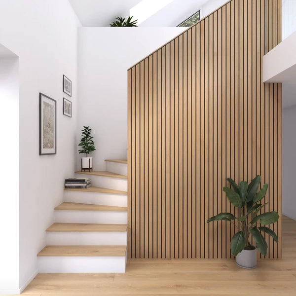 A well-lit interior scene displays a modern staircase with wooden steps and white risers leading to a landing with a small potted plant. Adjacent to the staircase, a vertical wooden slat wall extends upwards, creating a striking visual element alongside a larger potted plant in the foreground. Wall art adorns the white wall beside the stairs, and a skylight provides natural light from above.