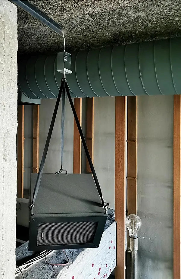 Hanging speaker with gray case and black grille suspended by black straps, alongside exposed ductwork and wooden framing.