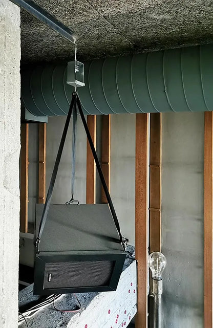 Hanging speaker with gray case and black grille suspended by black straps, alongside exposed ductwork and wooden framing.