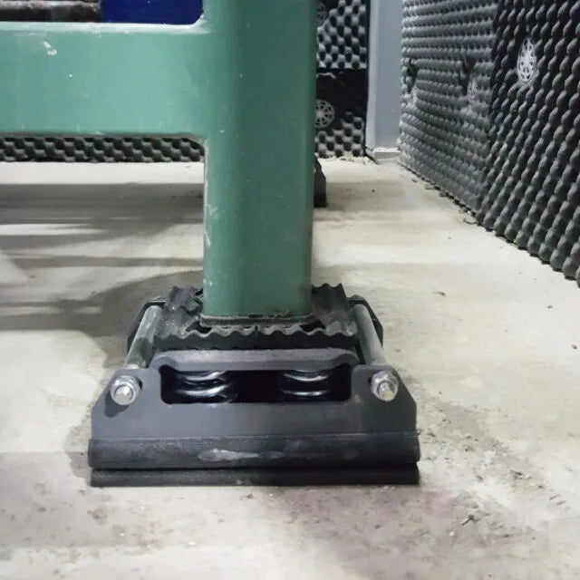 Close-up of a green metal furniture leg resting on a black vibration-dampening mount with visible springs, set on a concrete floor against a soundproof wall.