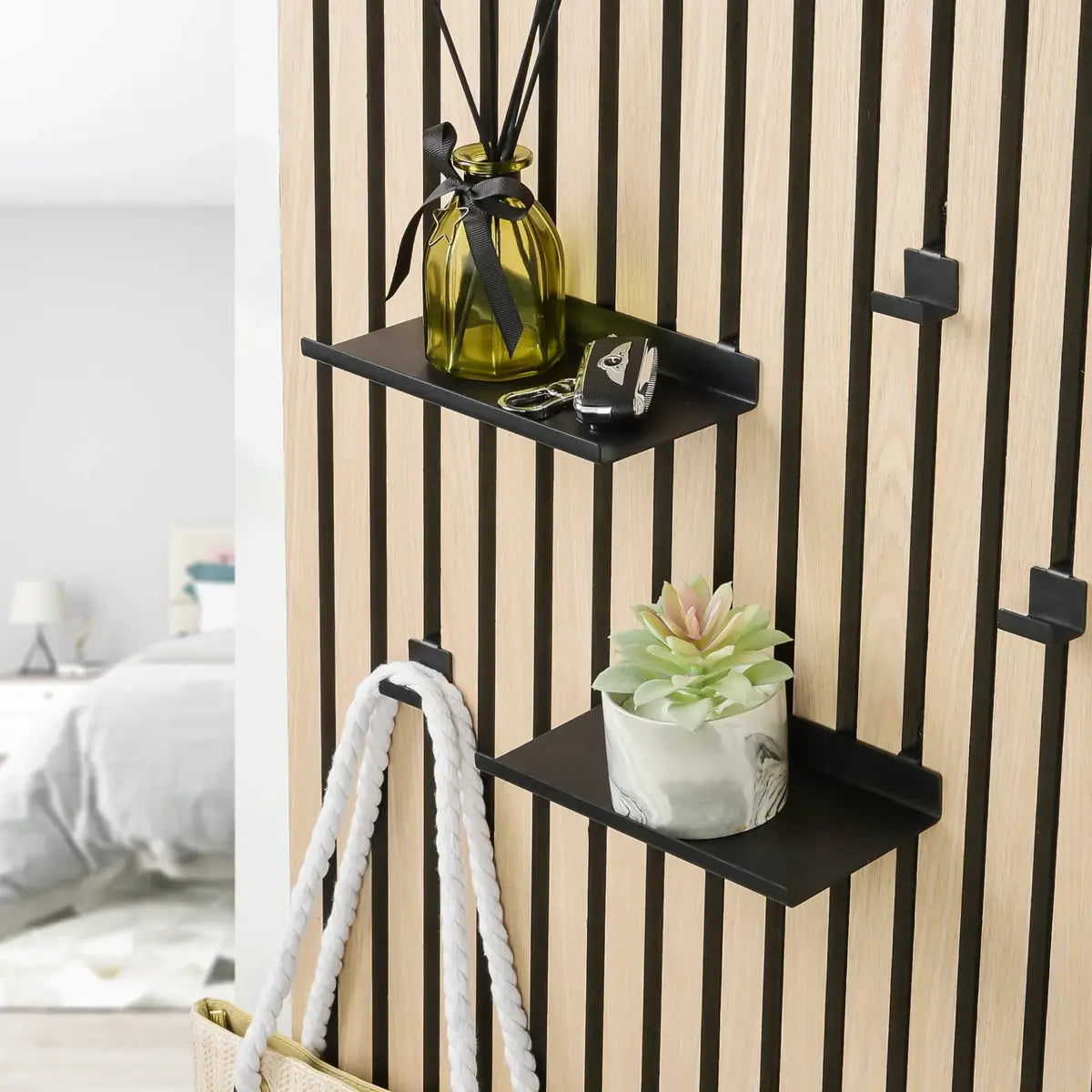 Close-up of two small black shelves attached to a vertically paneled wooden wall, displaying a glass bottle with diffuser sticks, keys, a potted succulent, and a white rope bag.
