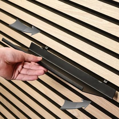 Close-up of a hand attaching a black metal shelf to a wooden slat wall with arrows indicating direction.
